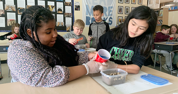 Two students holding a cup, pouring water into an experiment