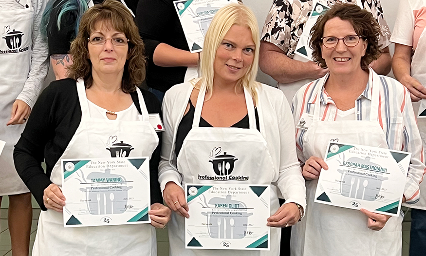 Three women in chef aprons hold certificates