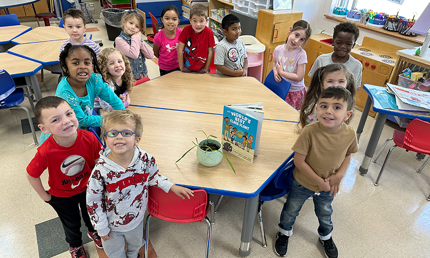 Kindergartners gather around table with plant and book on it
